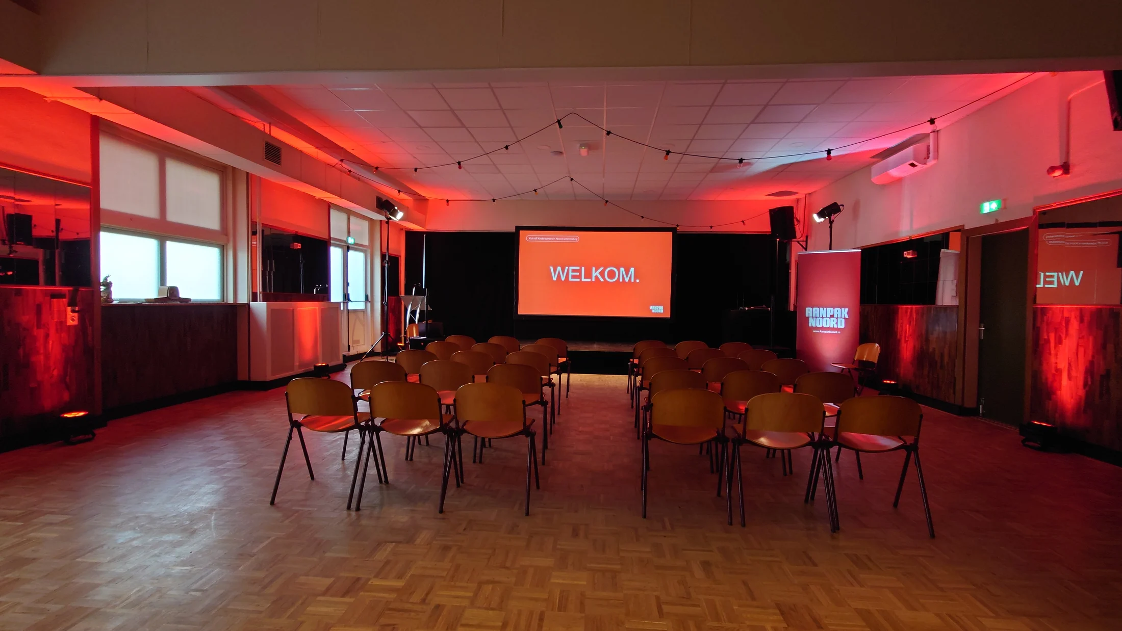 Zaalopstelling met podium, katheder, scherm en rode sfeerverlichting voor Aanpak Noord